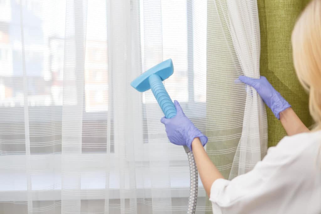 Femme portant des gants violets utilisant un nettoyeur vapeur pour assainir des rideaux blancs légers devant une fenêtre.